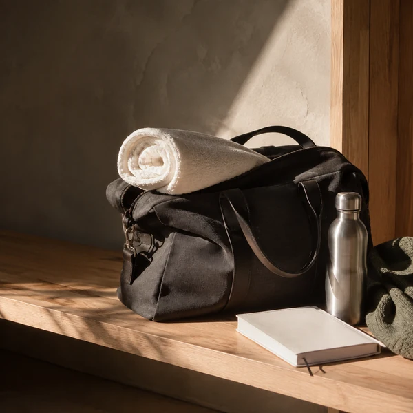 A black kit bag with a rolled towel, a steel water bottle and a notebook on a wooden bench